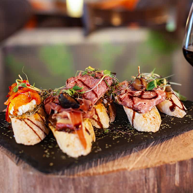 Montaditos Spanish tapa served on a slate platter sitting atop a slice of wood with a glass of red wine to its right